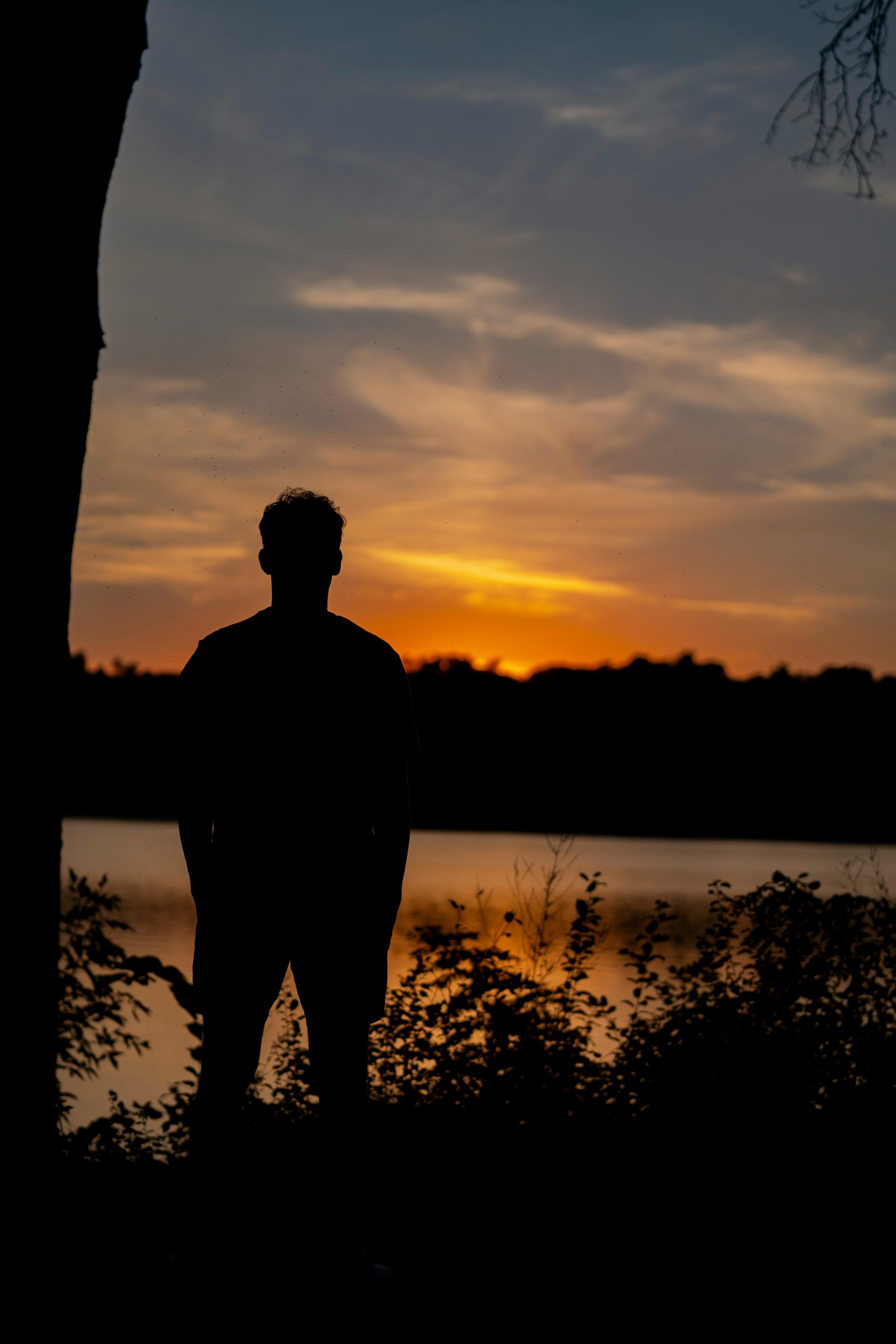 A lone silhouette stands by a serene lake observing a vibrant sunset.