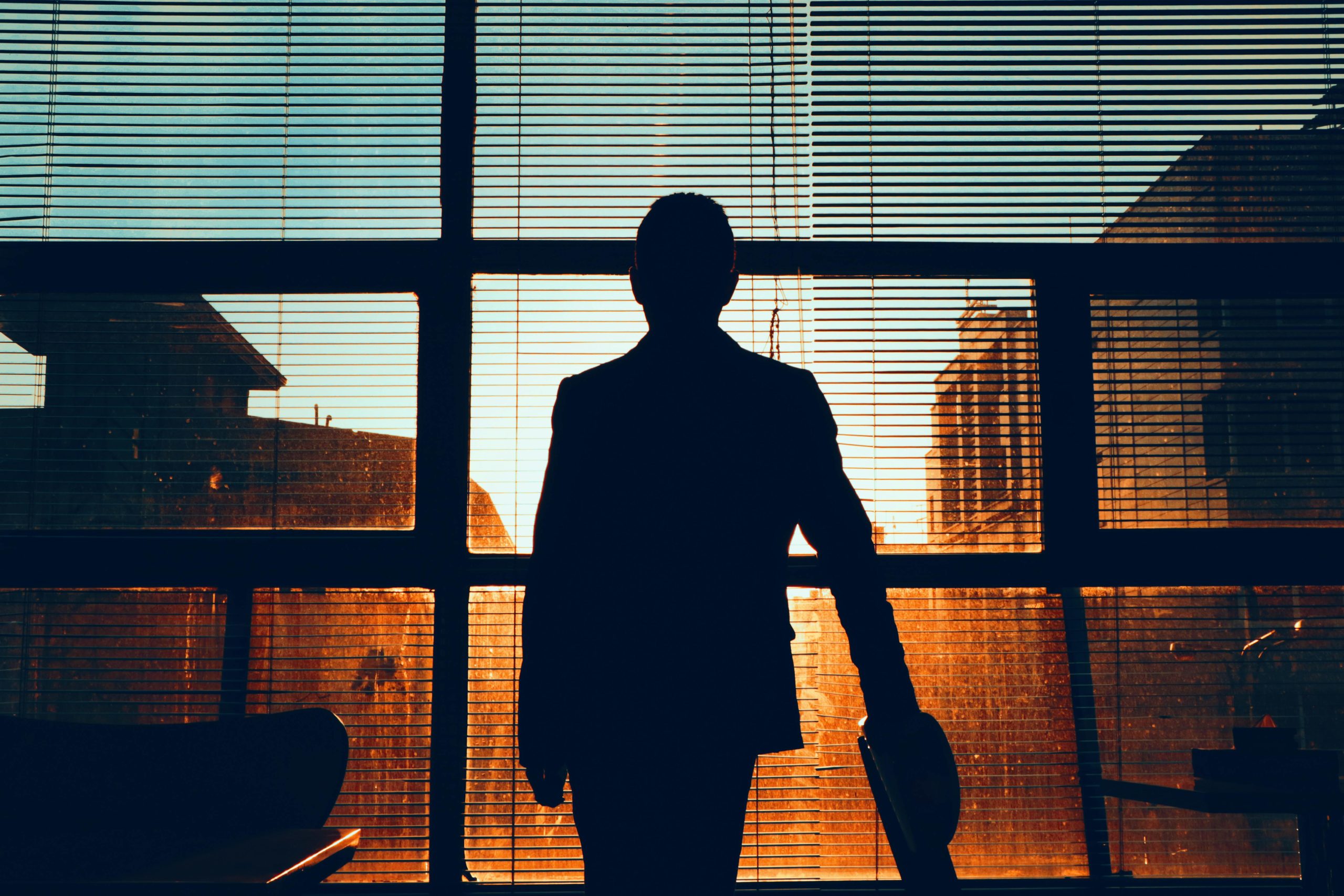Silhouette of a man standing by a window, observing a building at sunset.